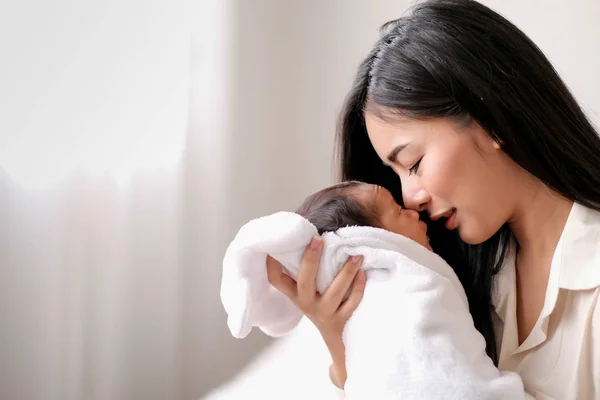 Beyaz gömlek Asyalı anne sevgi ve aile bağ göstermek için beyaz perde ile cam pencereler önünde yatak odasında onu yeni doğan bebek öpüyor.