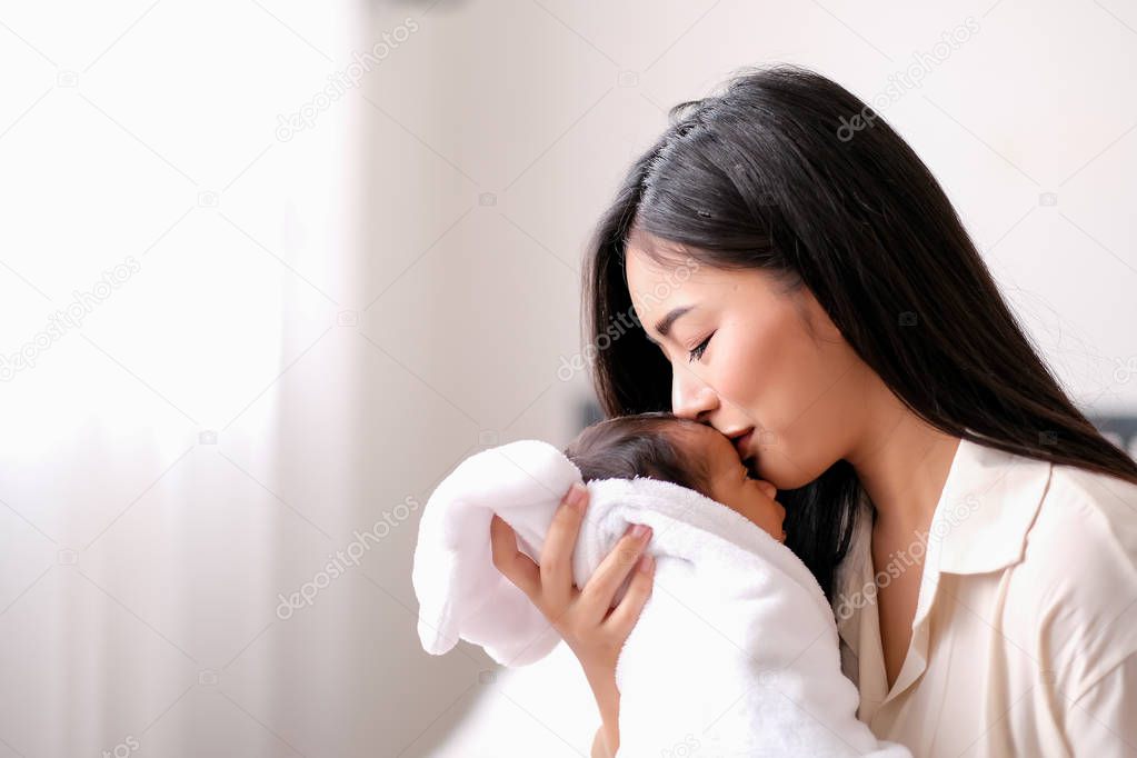 White shirt Asian mother is kissing at forehead of her newborn baby in bedroom in front of glass windows with white curtain to show love and family bonding.