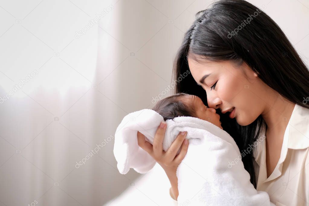White shirt Asian mother is kissing her newborn baby in bedroom in front of glass windows with white curtain to show love and family bonding.