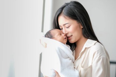 Beyaz gömlek Asyalı anne sevgi ve aile bağ göstermek için beyaz perde ile cam pencereler önünde yatak odasında onu yeni doğan bebek öpüyor.
