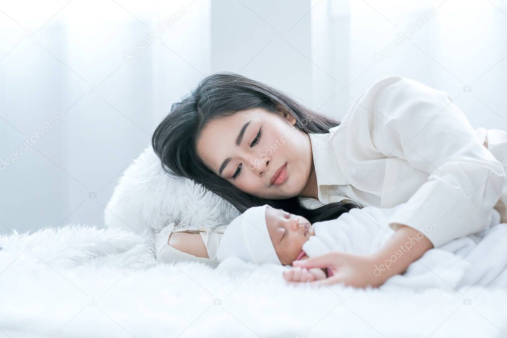 Asian mother lie on bed and look carefully to her newborn baby who sleep on white bed with day light.