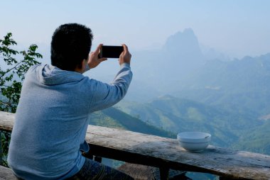Asyalı turist, güzel dağ resimlerini çekmek için cep telefonunu kullanıyor..