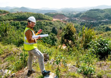 Beyaz miğferli mühendis müdürü ve inşaat kâğıdı planı ve el bilgisayarı ya da telsizi olan biri kameraya dönüp dağın önünde dursun..