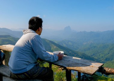 Asyalı bir turist bir tatil köyünün balkonunda kalıp kasede kahvaltı yapar ve mutlu ve rahat görünür..