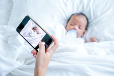 Anne, yeni doğan bebeğin beyaz yatakta uyurken fotoğrafını çekmek için cep telefonunu tutuyor..