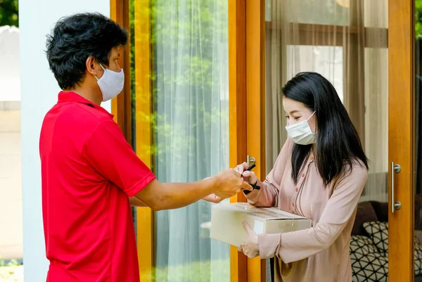 Beautiful woman with mask get delivery package box from carrier man with red shirt and mask then sign at home. Concept of new normal lifestyle to prevent virus infection during covid pandemic in city.