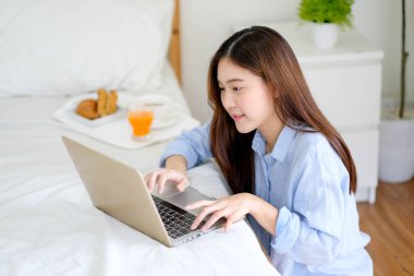 Güzel bir kadın çalışmak için dizüstü bilgisayar kullanır ve yatak odasında mutlu görünür ve ayrıca kahvaltıyı yatağa koyar. İş hayatında daha iyi sonuç elde etmek için evde çalışmanın mutluluk kavramı.