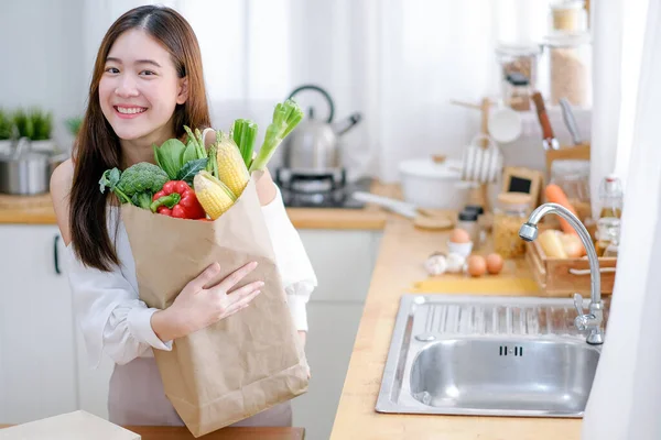 Lovely woman hold bag with various vegetables and smile stand in kitchen and look happy in morning light. Concept of healthy food for good and better health care management during work from home.