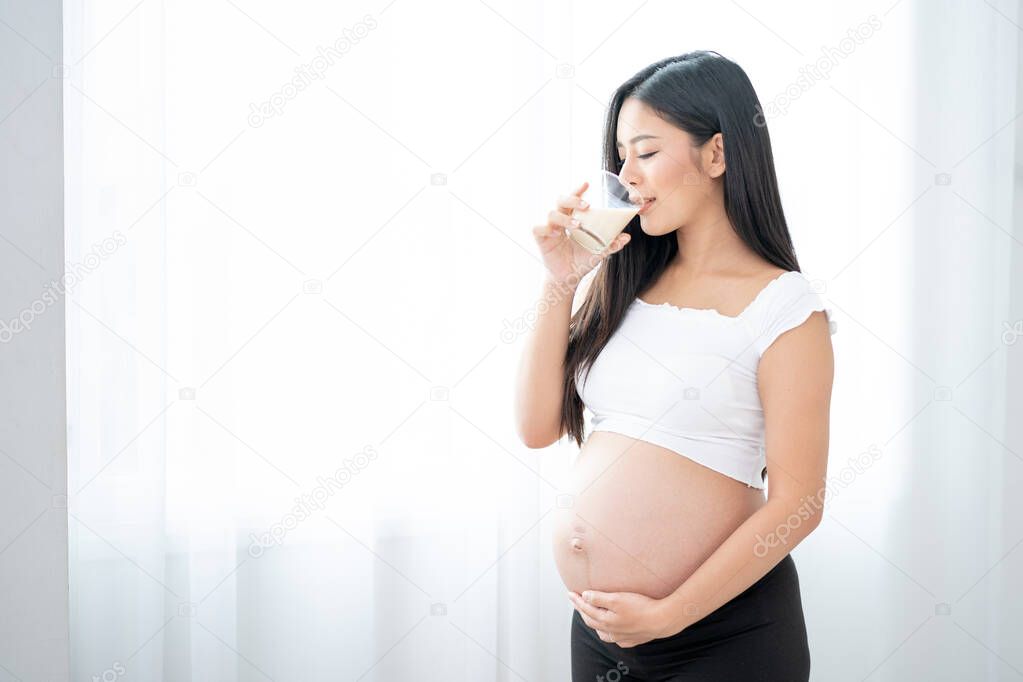 Close up beautiful Asian pregnant woman is drinking milk and stand in front of white curtain with morning light. Concept of good healthy food for mother and support baby in belly of people.