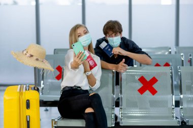 Maskeli birkaç beyaz erkek ve kadın, Covid-19 salgınından kurtulduktan sonra seyahat sırasında havalimanında sosyal mesafeli ve selfie ile birlikte oturuyorlar..