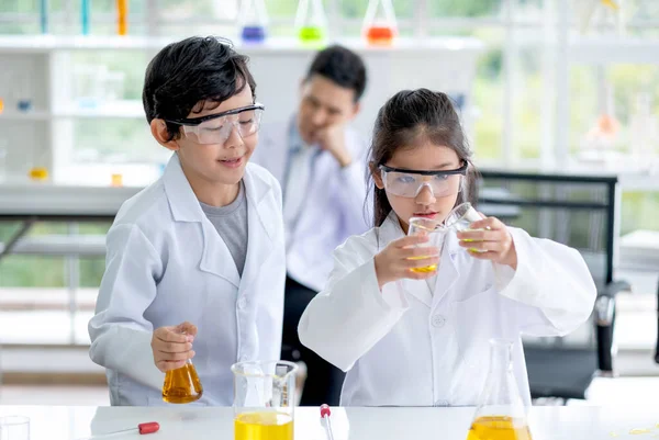 小男孩期待着小女孩在实验室的教室里倒一些彩色化学品来烧瓶 为儿童提供的良好做法和科学教育概念支持了他们的学习 — 图库照片