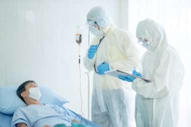 Tüm vücut örtüsü olan doktor hasta hastalığının ilerlemesini açıklıyor ve hemşire verileri Covid-19 hasta yatağının yanında kaydediyor..