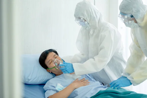 Nurse or hospital staff with full body cover suit help to put aspirator mask to Covid-19 infected patient who lie on bed in closed room.