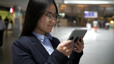Genç mutlu Asyalı iş kadını havaalanı koridorunda tablet kullanıyor.
