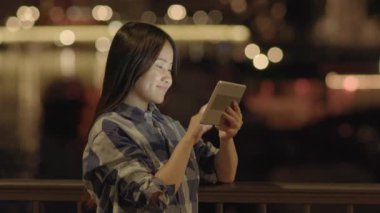Genç Asyalı kadın geceleri kentteki nehrin kıyısında dururken tablet kullanıyor. 