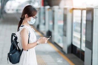 Surat maskesi takan genç Asyalı kız Coronavirus bükülmesini koruyor metro istasyonunda yürürken akıllı telefon kullanan mutlu turist kadın. yeni normal ve covid-19 salgınından sonraki yaşam