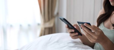 Kadın kredi kartı tutuyor ve cep telefonuyla online alışveriş yapıyor. Sabahları evde yatakta sipariş veriyor. İş, teknoloji, ekommerce, dijital bankacılık ve çevrimiçi ödeme kavramı
