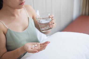 Yetişkin bir kadın elinde hap ve bir bardak su tutuyor, sabahları evde yatağında ilaç içiyor. Migren, ağrı kesici, baş ağrısı, grip, hastalık, hastalık ve sağlık sigortası kavramı