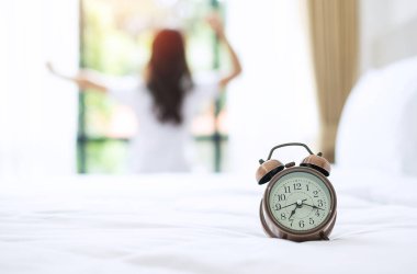 Uyandıktan sonra yatakta uzanan mutlu bir kadının arka planına sahip, kollarını kaldıran ve sabahları pencereye bakan genç yetişkin bir kadın. Rahatlayın ve güzel bir gün geçirin.