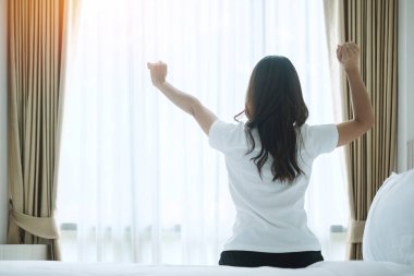 Uyandıktan sonra yatakta uzanan mutlu bir kadın, kollarını kaldıran ve sabahları pencereye bakan genç bir kadın. Rahatlayın ve güzel bir gün geçirin.
