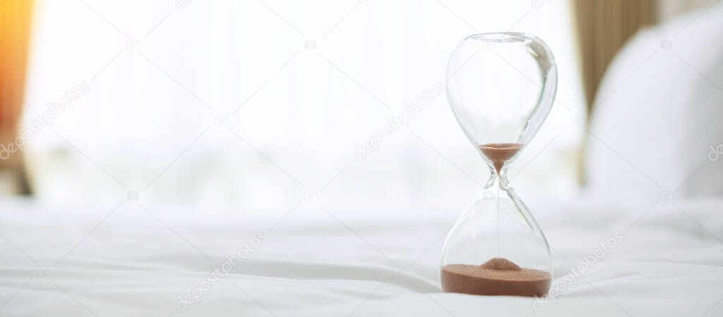 Hourglass on bed in the morning sunlight, Sand flowing through the bulb of Sandglass measuring the passing time. countdown, deadline, Life time and Retirement, daily routine concept