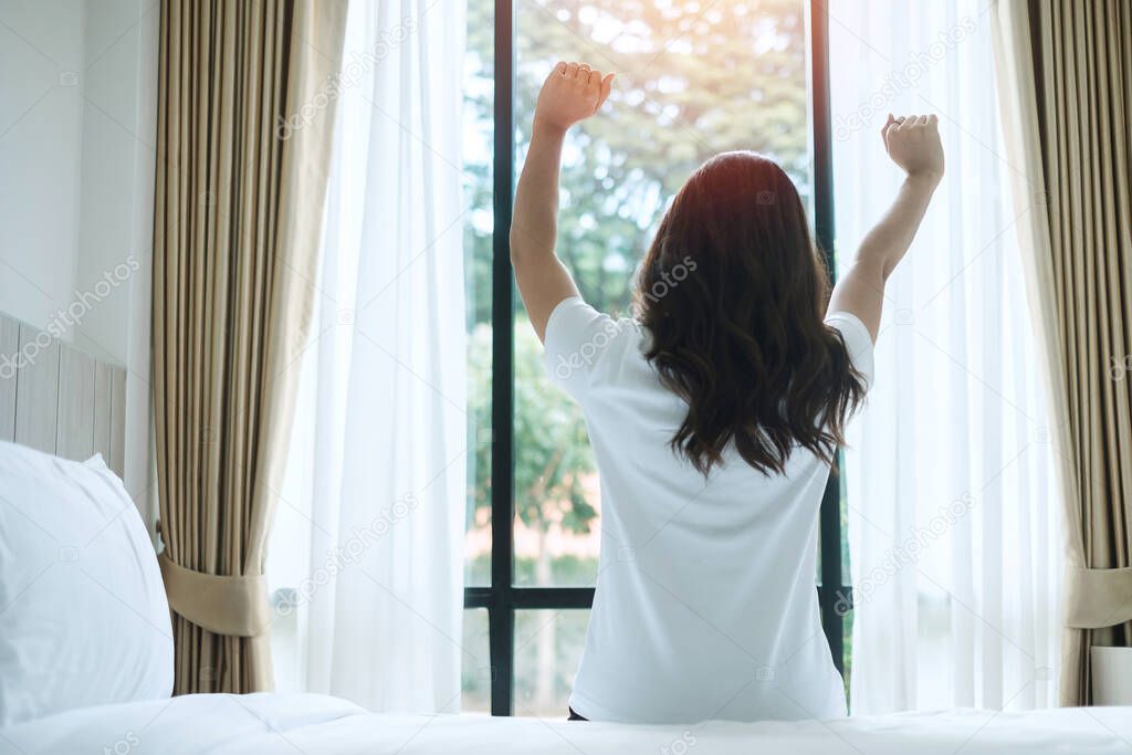 happy woman stretching in bed after waking up, young adult female rising arms and looking to window in the morning. fresh relax and have a nice day concepts