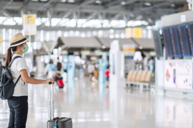 Havaalanında bagaj kontrolü yapan yüz maskesi takan genç bir kadın, Coronavirus hastalığı (Covid-19) enfeksiyonu, şapkalı Asyalı kadın gezgin. Yeni Normal ve seyahat balonu kavramı