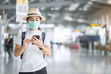 Havaalanı terminalinde yüz maskesi takan ve cep telefonu kullanan genç kadın, Coronavirus hastalığını (Covid-19) koruyan, şapkalı Asyalı kadın gezgin. Yeni Normal ve seyahat balonu kavramı