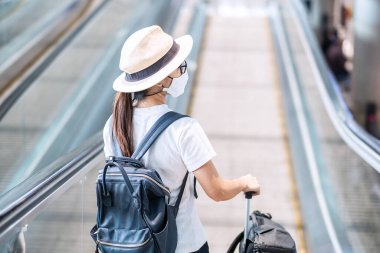 Havaalanı terminalinde yüz maskesi takan genç bir kadın, Coronavirus hastalığı enfeksiyonunu koruyan, havalimanında yürüyen merdivende yürüyen Asyalı bir kadın yolcu. Yeni Normal ve COVID-19 konsepti altında seyahat