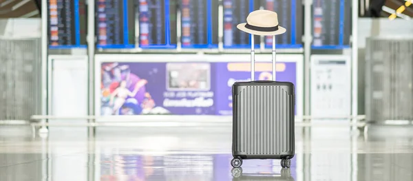 luggage bag with hat in international airport terminal, trolley suitcase with information board in aerodrome background. Transport, travel and vacation concepts