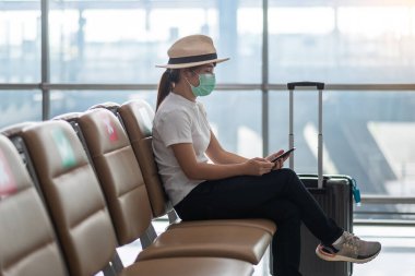 Havaalanında yüz maskesi takan ve cep telefonu kullanan genç bir kadın, Coronavirus enfeksiyonunu (Covid-19) koruma, sandalyede oturan Asyalı kadın yolcu. Yeni Normal ve Sosyal Mesafe