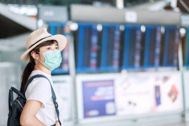 Havaalanı terminalinde cerrahi maske takan genç bir kadın, Coronavirus hastalığı (Covid-19) enfeksiyonunu koruyan, şapkalı Asyalı gezgin bir kadın. Yeni Normal ve seyahat balonu kavramı