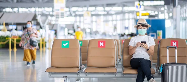 Young female wearing face mask and using mobile smartphone in airport, protection Coronavirus disease (Covid-19) infection, Asian woman traveler sitting on chair. New Normal and social distancing