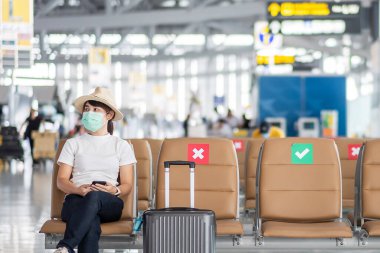 Yüz maskesi takan ve havaalanında sandalyeye oturan genç bir kadın, Coronavirus hastalığı (Covid-19) enfeksiyonu, Asyalı kadın gezgin. Yeni Normal, seyahat balonu ve sosyal uzaklık