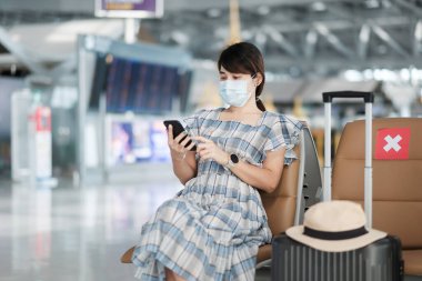 Havaalanında yüz maskesi takan ve cep telefonu kullanan genç bir kadın, Coronavirus enfeksiyonunu (Covid-19) koruma, sandalyede oturan Asyalı kadın yolcu. Yeni Normal ve Sosyal Mesafe