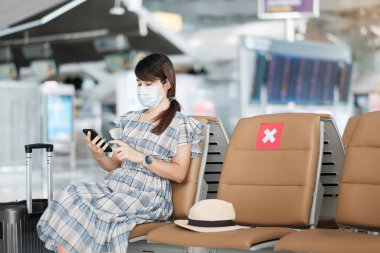 Havaalanında yüz maskesi takan ve cep telefonu kullanan genç bir kadın, Coronavirus enfeksiyonunu (Covid-19) koruma, sandalyede oturan Asyalı kadın yolcu. Yeni Normal ve Sosyal Mesafe