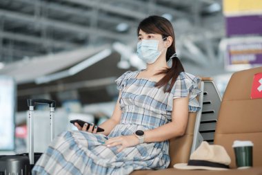 Havaalanında yüz maskesi takan ve cep telefonu kullanan genç bir kadın, Coronavirus enfeksiyonunu (Covid-19) koruma, sandalyede oturan Asyalı kadın yolcu. Yeni Normal ve Sosyal Mesafe