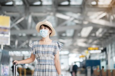 Bavul taşıyan yüz maskesi takan genç bir kadın havaalanında uçuş süresini kontrol ediyor, Coronavirus hastalığı enfeksiyonunu koruyor, seyahat etmeye hazır Asyalı bir kadın yolcu. Yeni Normal ve COVID-19 altında seyahat 