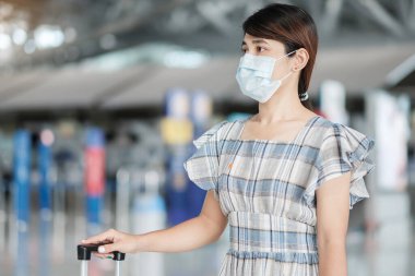Bavul taşıyan yüz maskesi takan genç bir kadın havaalanında uçuş süresini kontrol ediyor, Coronavirus hastalığı enfeksiyonunu koruyor, seyahat etmeye hazır Asyalı bir kadın yolcu. Yeni Normal ve COVID-19 altında seyahat 