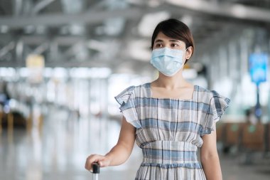 Bavul taşıyan yüz maskesi takan genç bir kadın havaalanında uçuş süresini kontrol ediyor, Coronavirus hastalığı enfeksiyonunu koruyor, seyahat etmeye hazır Asyalı bir kadın yolcu. Yeni Normal ve COVID-19 altında seyahat 