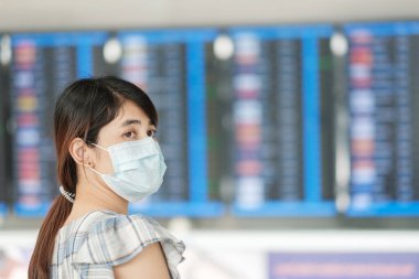 Cerrahi maske takan genç bir kadın havaalanında uçuş süresini kontrol ediyor, Coronavirus hastalığı enfeksiyonunu koruyor, seyahat etmeye hazır Asyalı bir kadın yolcu. COVID-19 altında Yeni Normal ve güvenli seyahat