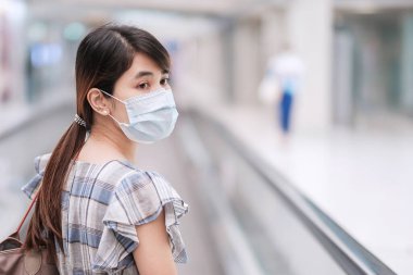 Havaalanı terminalinde yüz maskesi takan genç bir kadın, Coronavirus hastalığı enfeksiyonunu koruyan, havalimanında yürüyen merdivende yürüyen Asyalı bir kadın yolcu. Yeni Normal ve COVID-19 konsepti altında seyahat