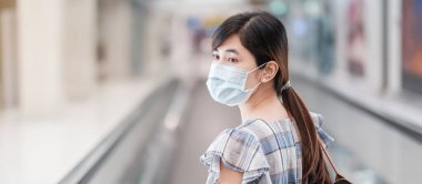 Havaalanı terminalinde yüz maskesi takan genç bir kadın, Coronavirus hastalığı enfeksiyonunu koruyan, havalimanında yürüyen merdivende yürüyen Asyalı bir kadın yolcu. Yeni Normal ve COVID-19 konsepti altında seyahat
