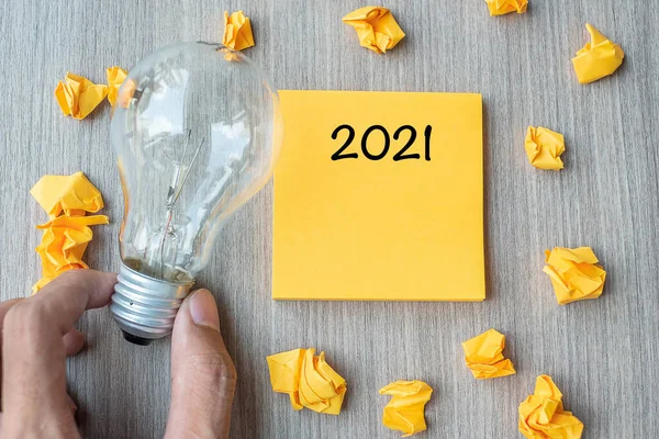 2021  words on yellow note and crumbled paper with Businessman holding lightbulb on wooden table background. New Year New Idea Creative, Innovation, Imagination, Resolution and Goal concept