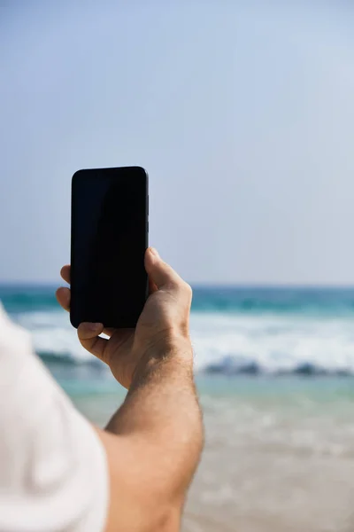 Cellulare in mano maschile sullo sfondo della spiaggia e dell'oceano . Fotografia Stock