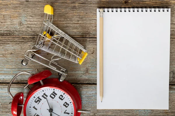 Shopping time concept. Shopping cart and a stopwatch on an old wood background with a copy space for shopping or sale offer list. Pencil and notepad with a place for text