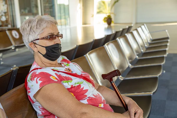 Havaalanının bekleme salonunda el yapımı yüz maskesi takmış yaşlı bir kadın oturuyor. Coronavirüs salgını konseptine bağlı seyahat kısıtlamaları