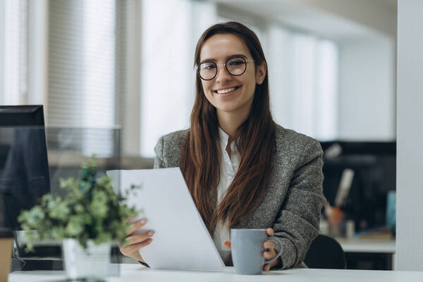 Portrait of smiling pretty young business woman in glasses sitting on workplace and working with documents.