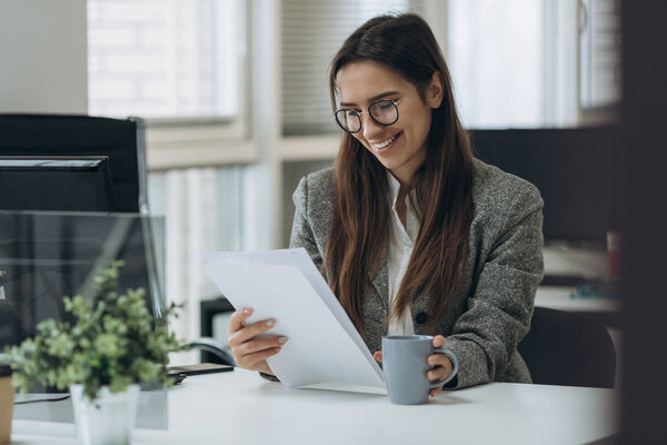 Portrait of smiling pretty young business woman in glasses sitting on workplace and working with documents.