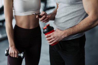 Fitness, spor, teknoloji ve zayıflama kavramı - kadar yakın genç kadın ve akıllı telefon ve su şişeleri içinde jimnastik salonu ile kişisel eğitmen gülümseyen